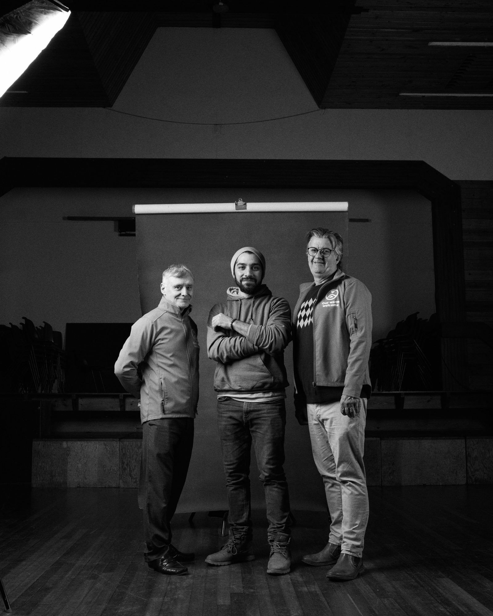 Abdullrhman Hassona stands with two supporters of the project in a large wooden floored room, the image is in black and white.