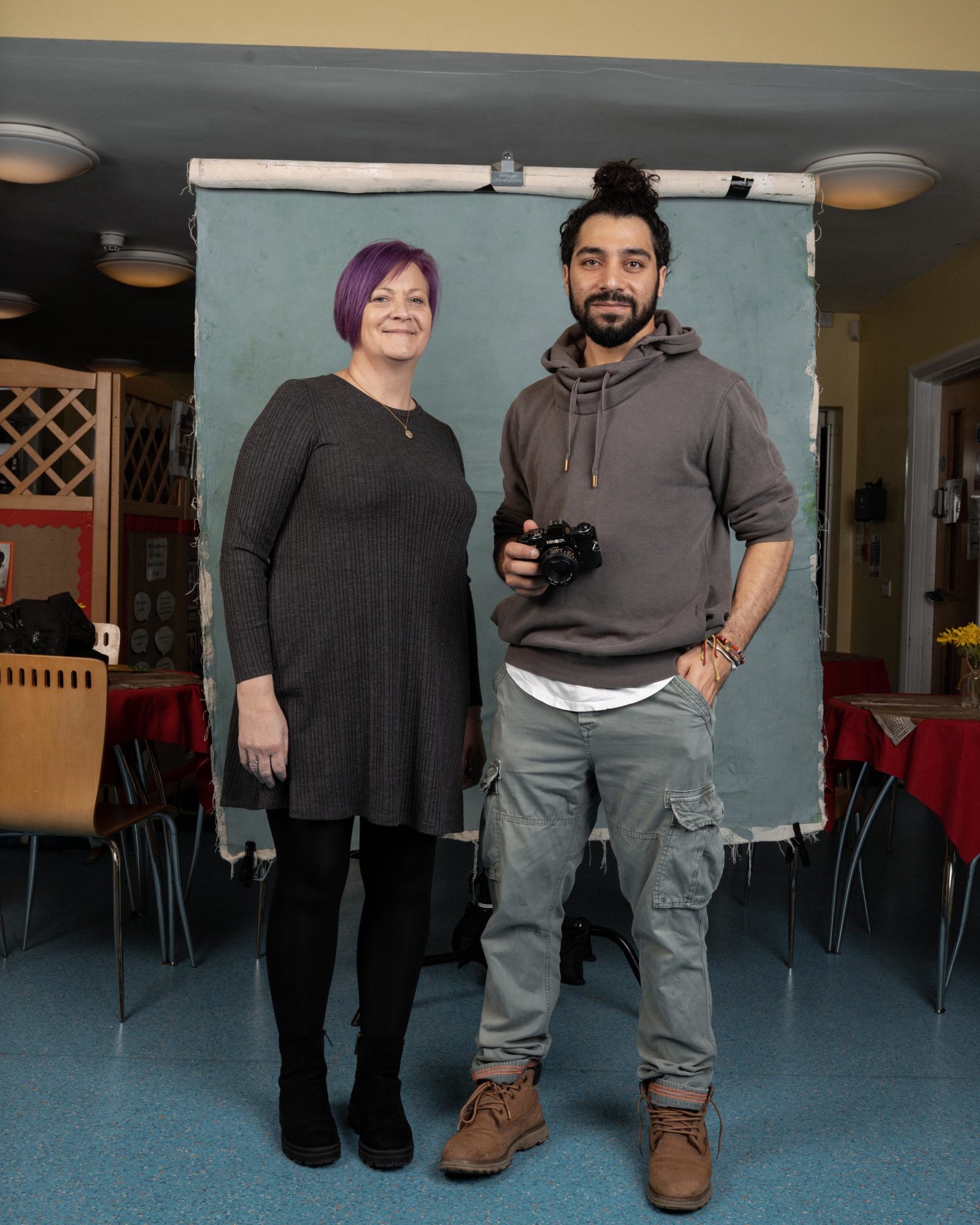 A woman with purple hair stands next to a man on a photography set in front of a screen