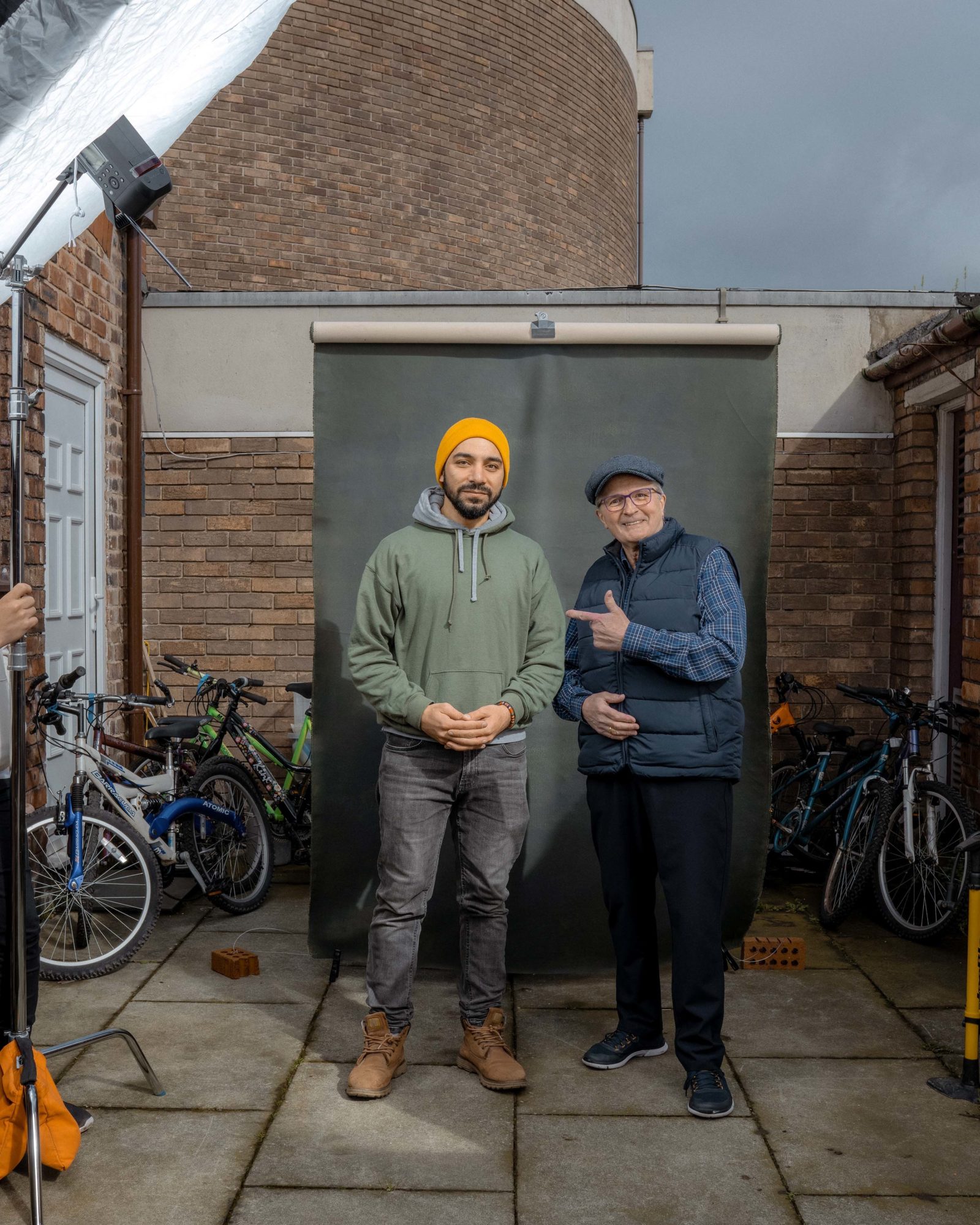 Two men stand outside in a yard in front of a photography screen, surrounded by bikes
