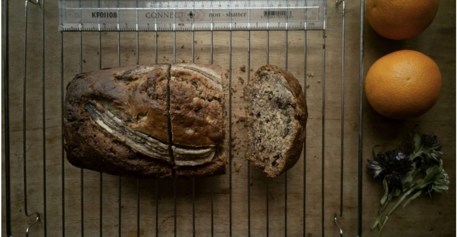 Image of a loaf of bread on a cooling rack from birds eye view, there is a ruler above it on the rack and the bread has 2 large chunks sliced off the end. There are two oranges and some herbs beside the rack.