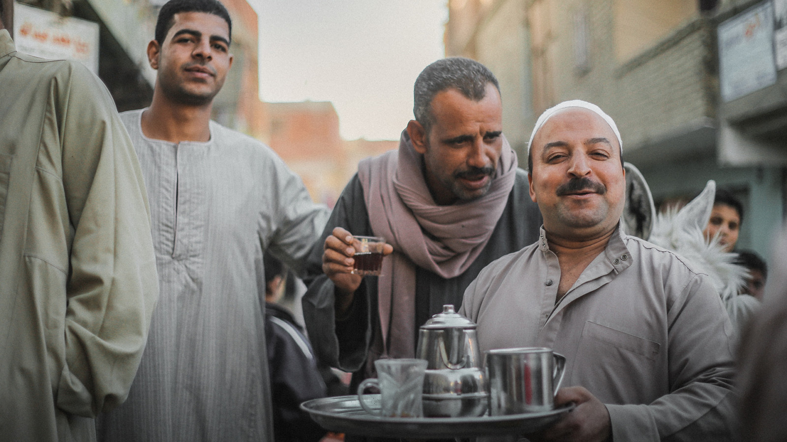 3 men in a crowded street, the man in the foreground is smiling and holding a tray with a silver teapot, a silver jug and a glass mug on it, there is a man holding a glass of what appears to be black tea or coffee and is saying something to the other man. There is a third man in the background looking at the camera candidly.
