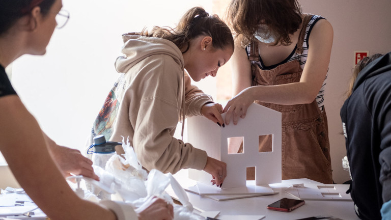Two young women build a model of a house together from cardboard