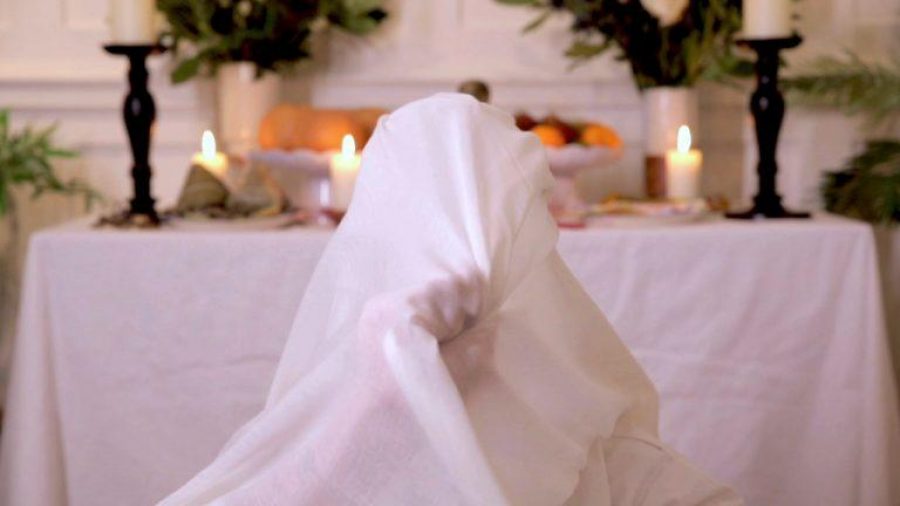 A person wrapped in a white sheet holding on from the inside so we can see the shape of their fist gathering the material. Behind them a table covered in a white sheet has lit pillar candles, candle stands, a large bowl of fruit and foliage arranged like an altar.