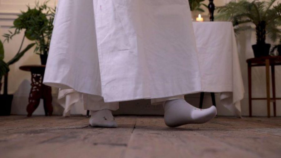 A pair of feet wearing white socks underneath a white gown and trousers. They are standing on wooden floor and behind them a table with a white sheet and candles on top, and lots of green foliage.