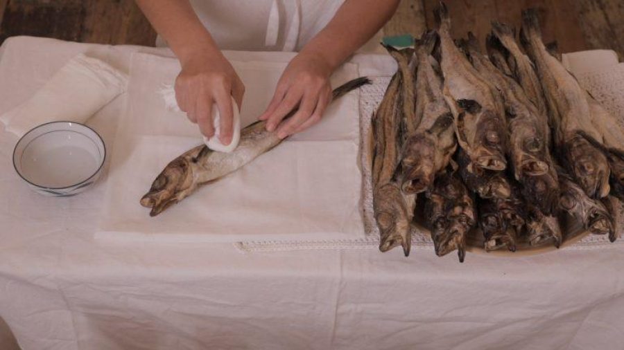 On a table with a white cloth on a pair of hands clean a dried fish with a bowl of water and a white cloth. Next to them a large pile of dried fish.