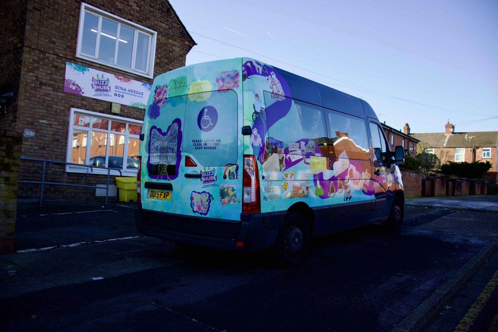 Buzz Hub bus from the back with Rudy Loewe's vinyl wrap applied, the bus is dark blue and the wrap has a turquoise background with a purple wiggly line wrapping around it and the workshop illustrations dotted around it.