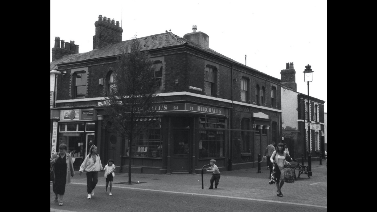 A black and white photograph of a high street with people walking.