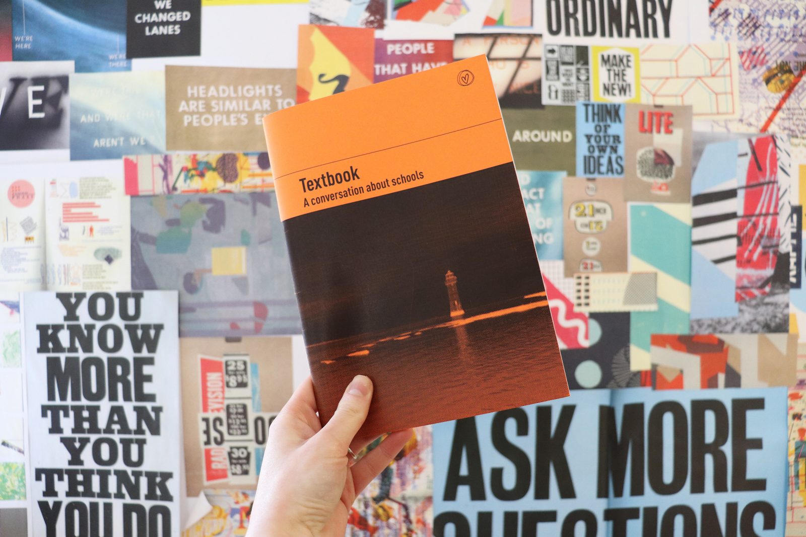 A hand holds a black and orange booklet up in front of a wall full of posters. On the front of the book is an image of a lighthouse and the title 'Textbook'.