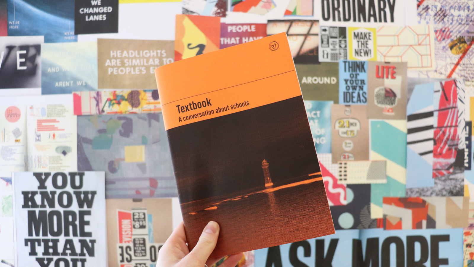A hand holds a black and orange booklet up in front of a wall full of posters. On the front of the book is an image of a lighthouse and the title 'Textbook'.