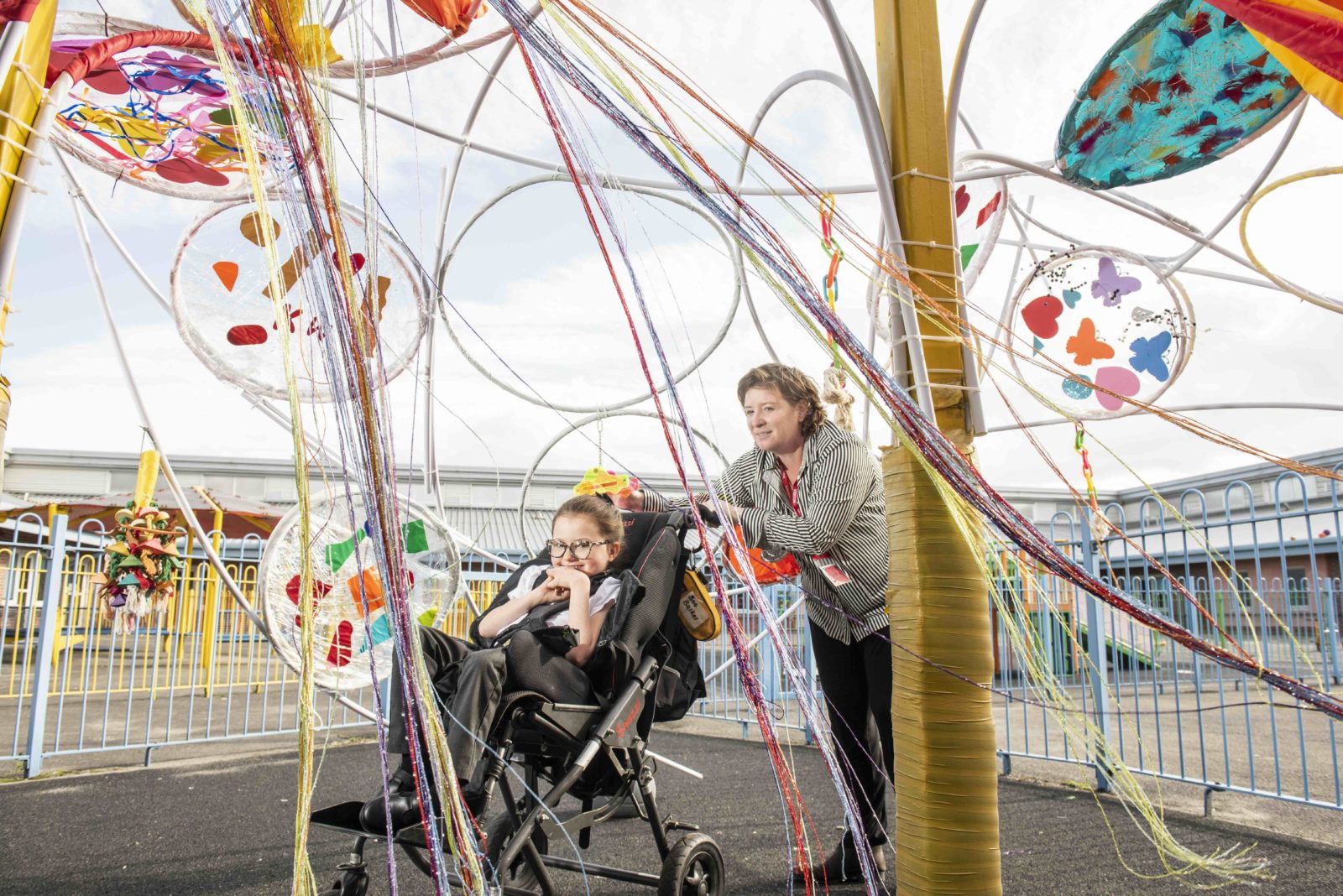 A woman in a stripy shirt is pushing a small girl in a wheelchair underneath a brightly coloured artwork made of streamers and hula hoops in a playground. The woman is smiling and the young girl is grinning.