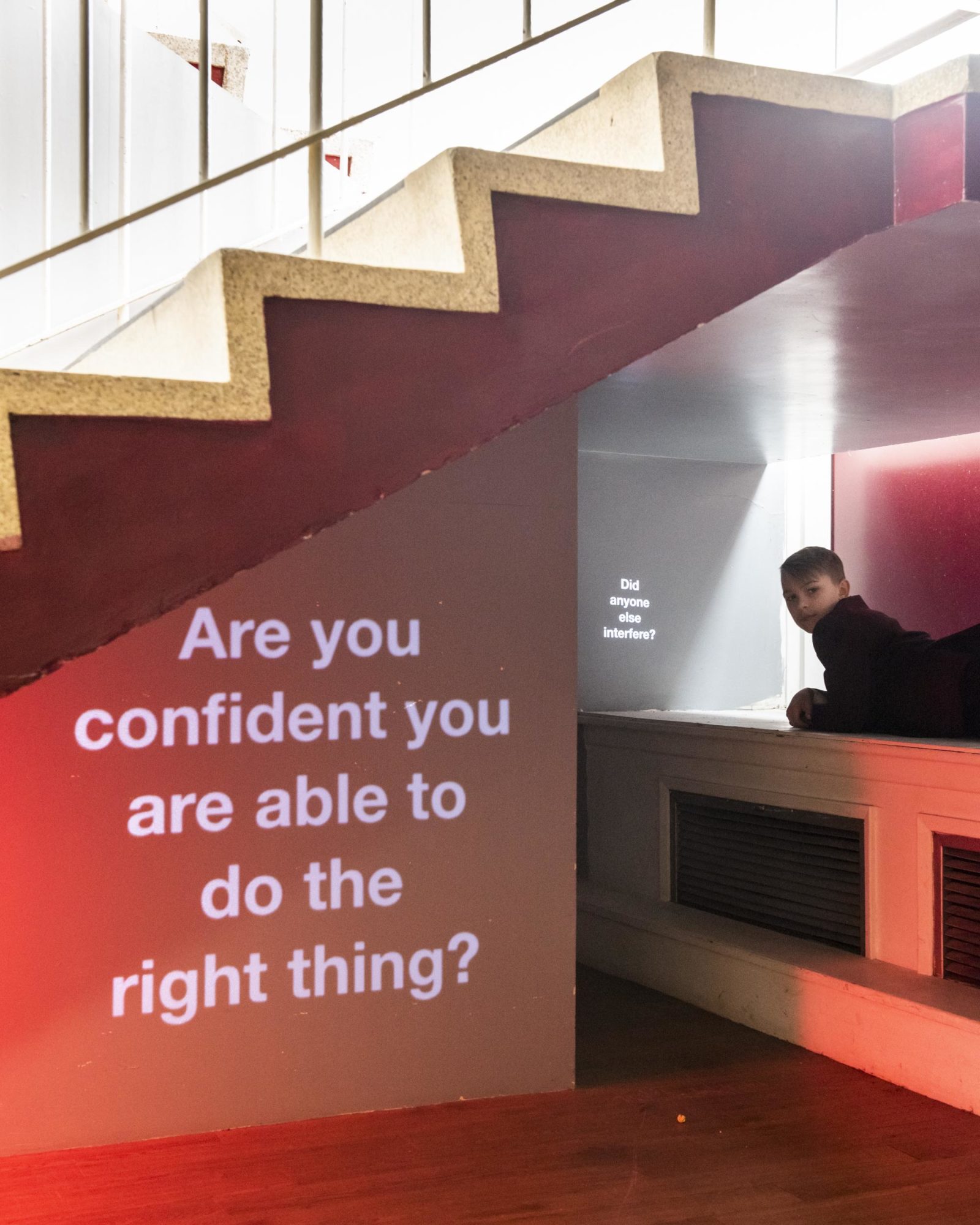 In a room with white walls, desk and stairs and a red carpet the words 'Are you confident you are able to do the right thing?' are projected on to a wall.
