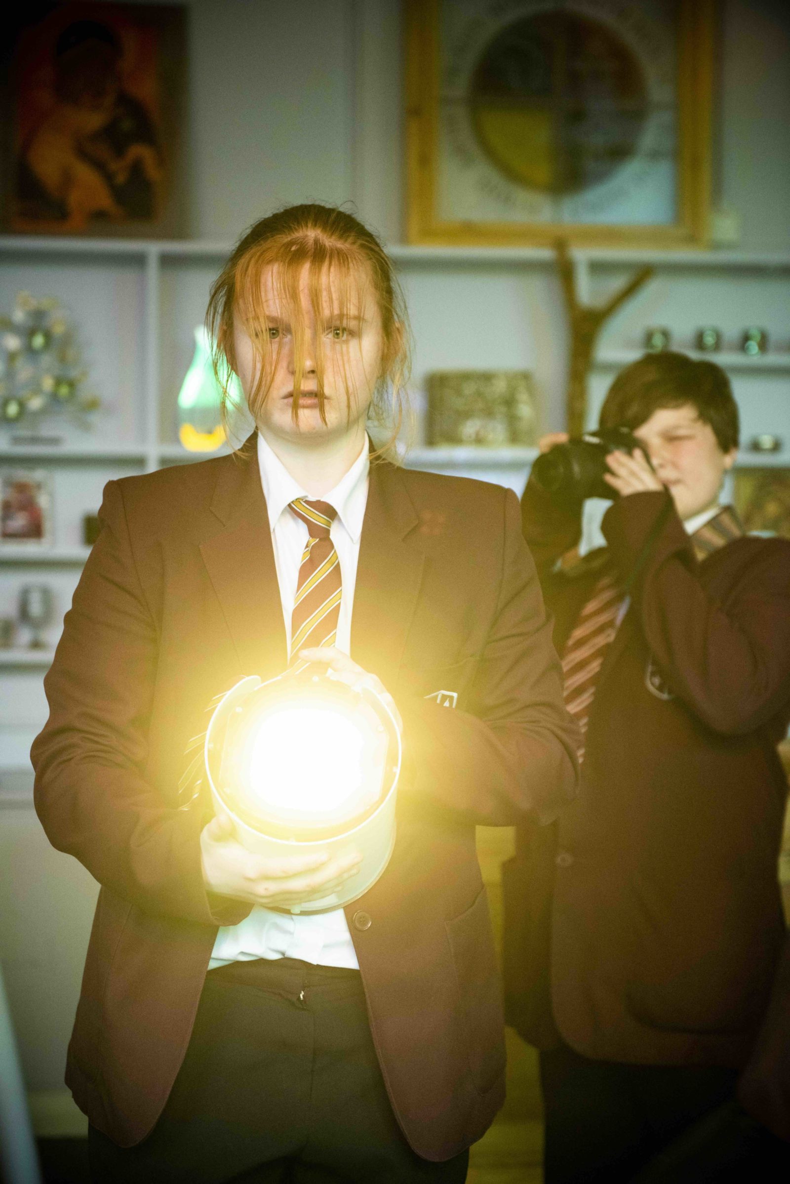 A young person in a school uniform is facing the camera, holding a large spotlight and shining towards us. They have long red hair which falls over their eyes and serious look on their face. Behind them another young person in a school uniform is holding up a camera and taking a picture.