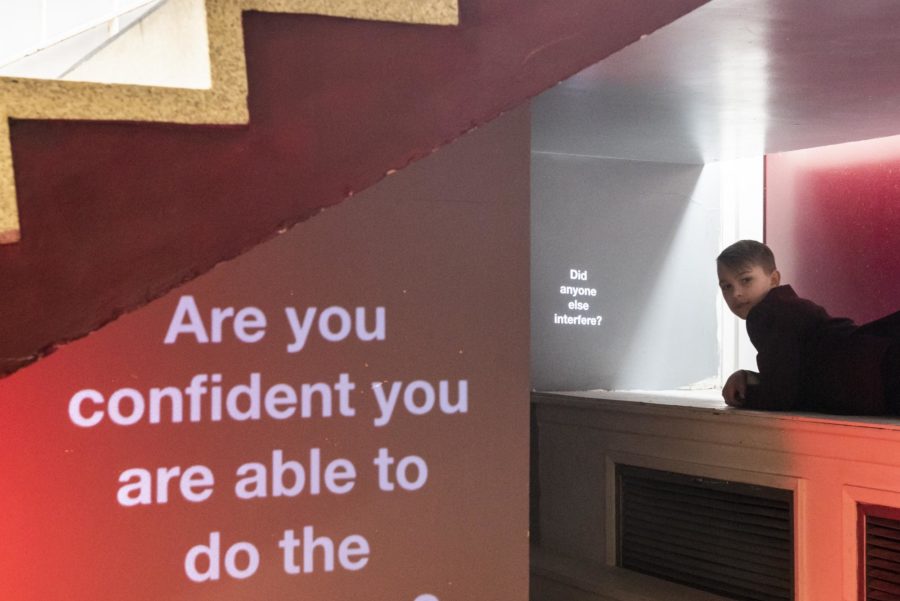 In a room with white walls, desk and stairs and a red carpet the words 'Are you confident you are able to do the right thing?' are projected on to a wall.