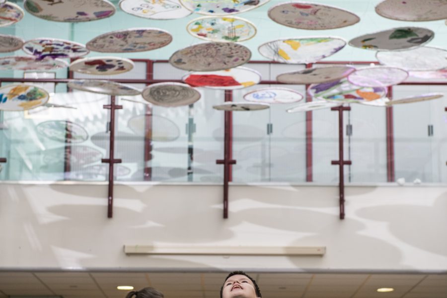 A canopy of circular artworks hangs above a room. Some of the circles are plain white, some have abstract, colourful designs. A young person stands underneath looking up towards the artworks. Some people are gathered in the background.