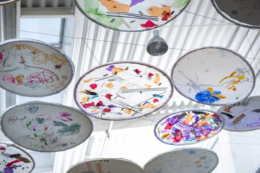 A canopy of circular artworks hang underneath a plastic ceiling. The artworks are colourful and abstract with light shining through from the ceiling.