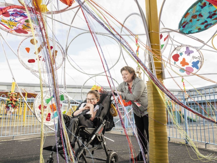 A woman in a stripy shirt is pushing a small girl in a wheelchair underneath a brightly coloured artwork made of streamers and hula hoops in a playground. The woman is smiling and the young girl is grinning.