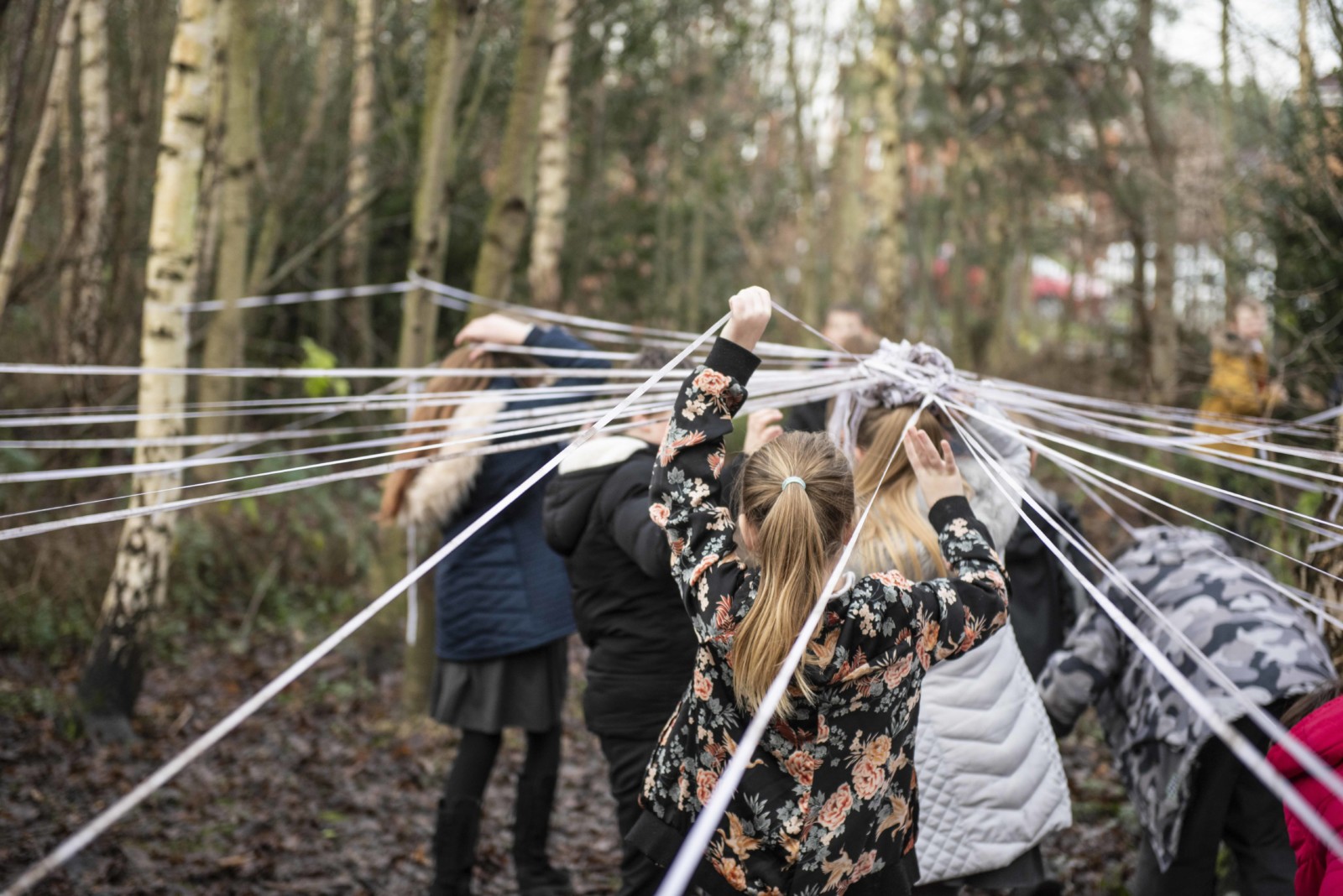 Children walk through the forest hiding up white ribbons that are wrapped around the tree trunks