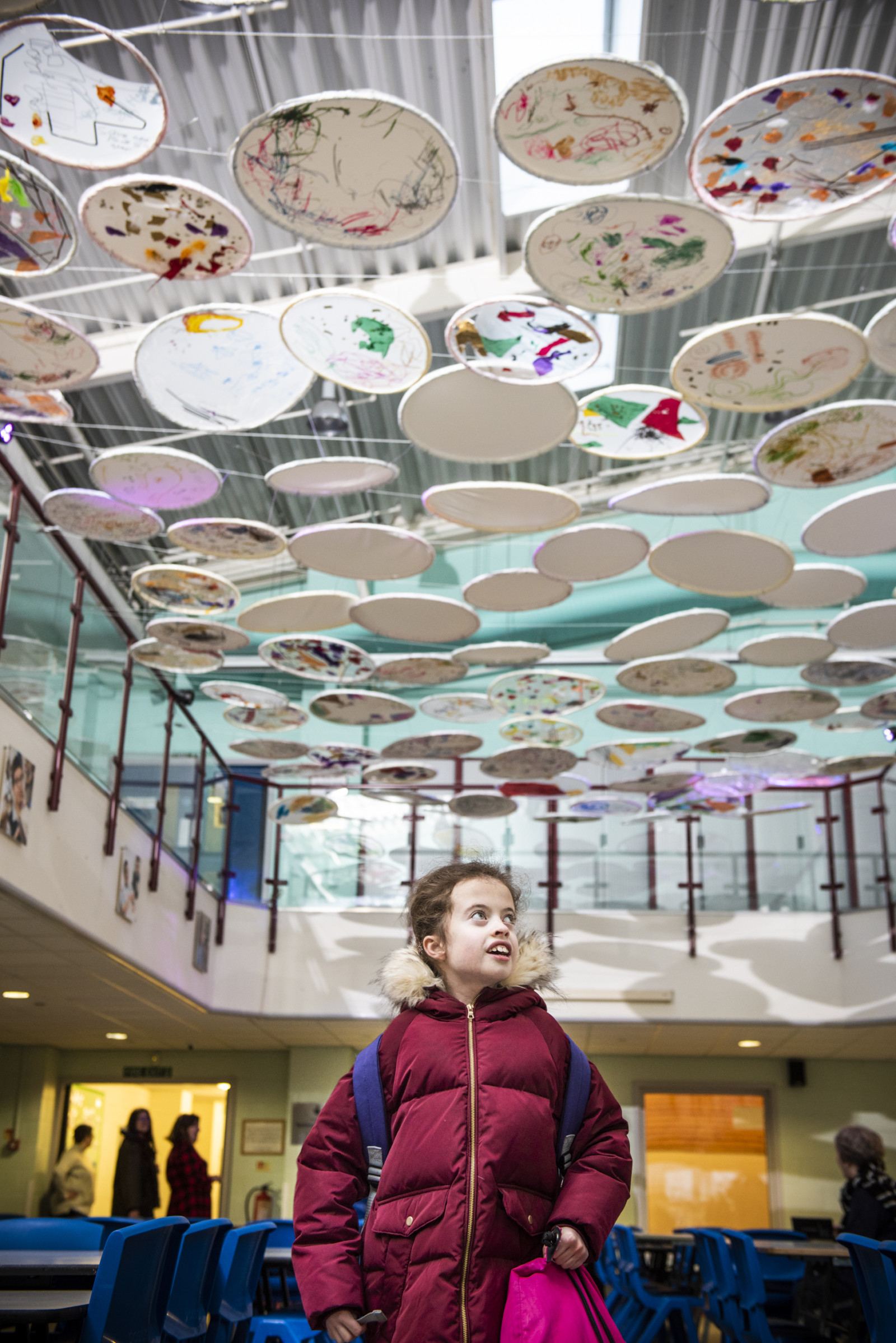 A young girl in a pink coat looks up to an installation hanging over her. The installation consists of many seemingly floating and decorated disks attached together.