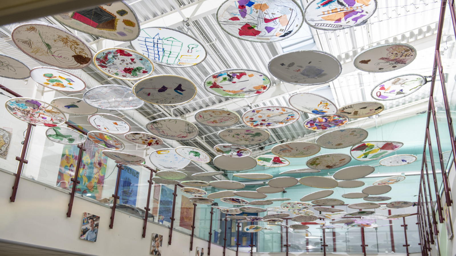 In a large bright room a collection of circles are suspended from one balcony to another, above a seating area. The circles are decorated with patterns in many different colours and shapes.