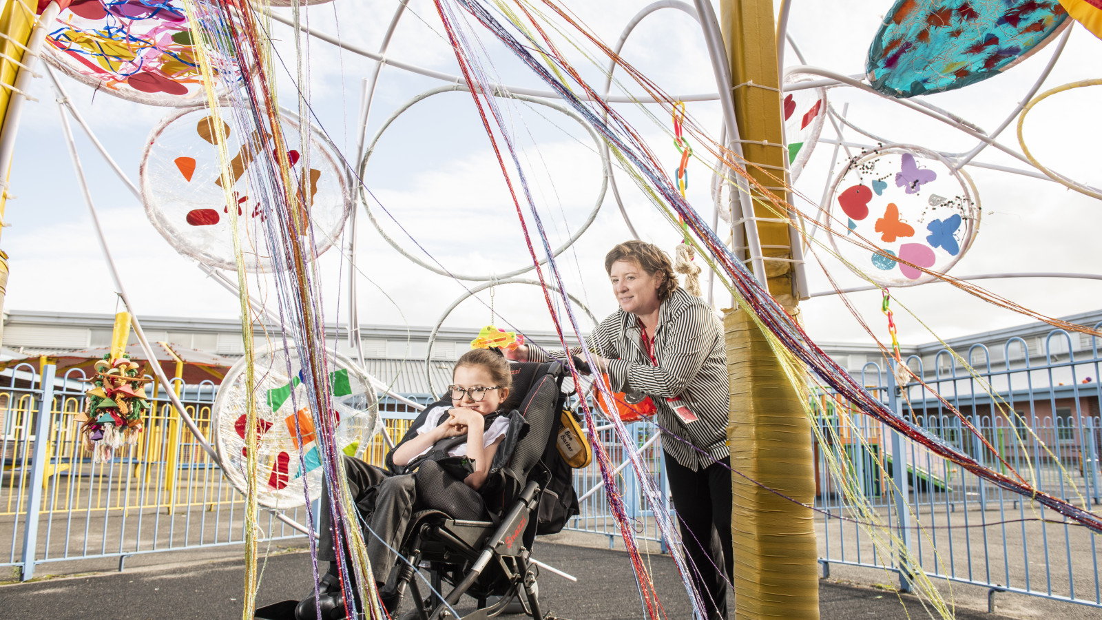 A woman in a stripy shirt is pushing a small girl in a wheelchair underneath a brightly coloured artwork made of streamers and hula hoops in a playground. The woman is smiling and the young girl is grinning.