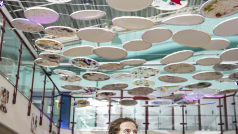 In a large bright room a collection of circles are suspended from one balcony to another, above a seating area. The circles are decorated with patterns in many different colours and shapes. A child walks underneath the suspended circles, gazing upwards and smiling.