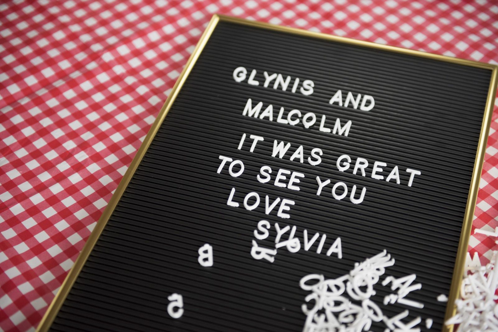 On a red and white checked background a black pin board rests with white letters on. The letters read 'Glynis and Malcolm it was great to see you love sylvia' A jumble of white letters are piled at the bottom of the pin board.