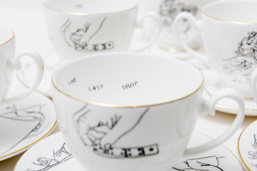 A set of 5 white teacups and saucers with gold rim and black line drawing design. Inside one of the teacups you can see the words 'the last drop' in black.