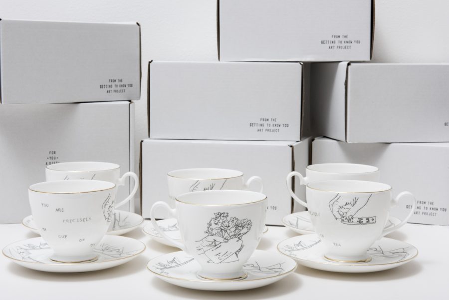 A set of white teacups and saucers sit in front of white boxes. The teacups are decorated with line drawings of hands playing dominoes, flowers with tags attached and some text which reads 'you are precisely my cup of tea'. A black stamp on the boxes reads 'from the getting to know you art project'