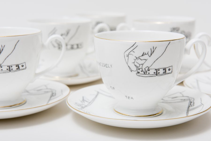 Five teacups and saucers against a white background. The teacups are white with a golden rim, and show a black line drawing design of some hands playing dominoes.