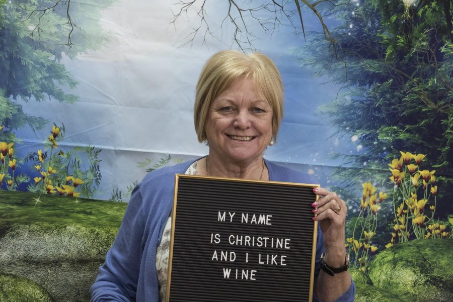 A person with fair hair in a bob sits in front of a colourful backdrop holding a black pin board. The backdrop is a forest design, with trees, mossy and red and white toadstools. The pin board they hold reads 'My name is Christine and I like Wine.'