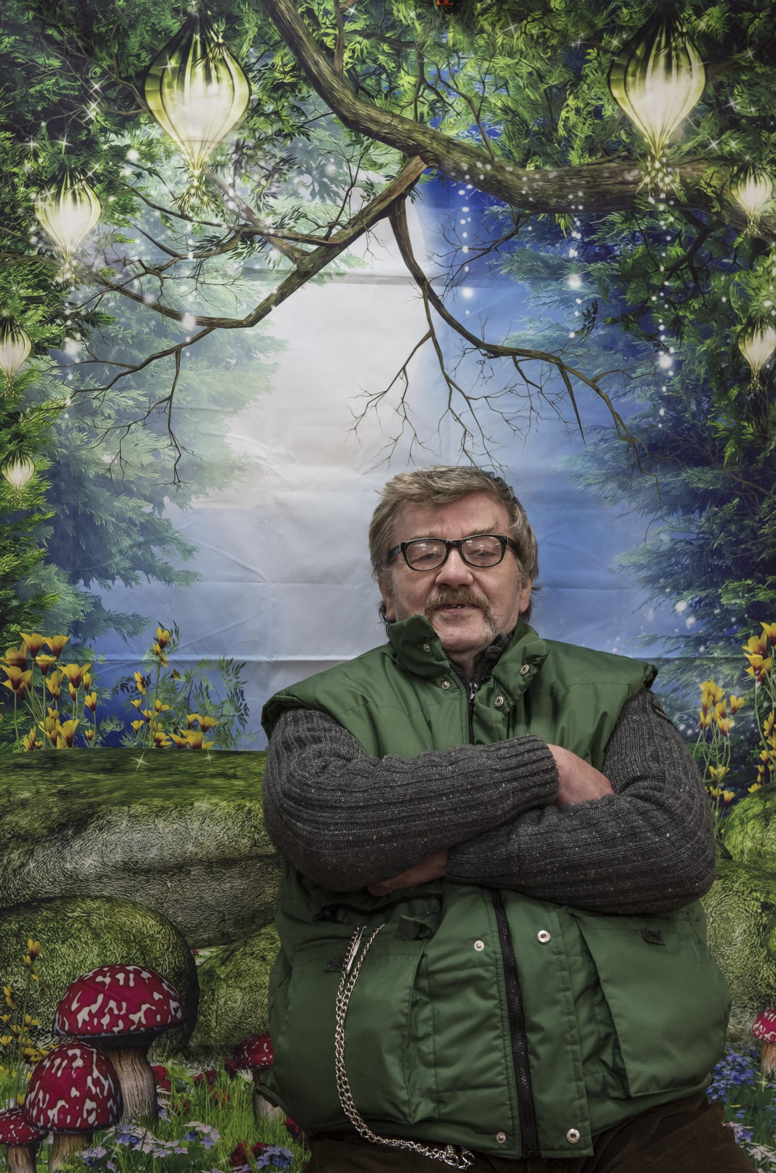 A person with glasses and short dark hair sits with their arms crossed in front of a colourful backdrop. The backdrop is a forest design, with trees, mossy and red and white toadstools.