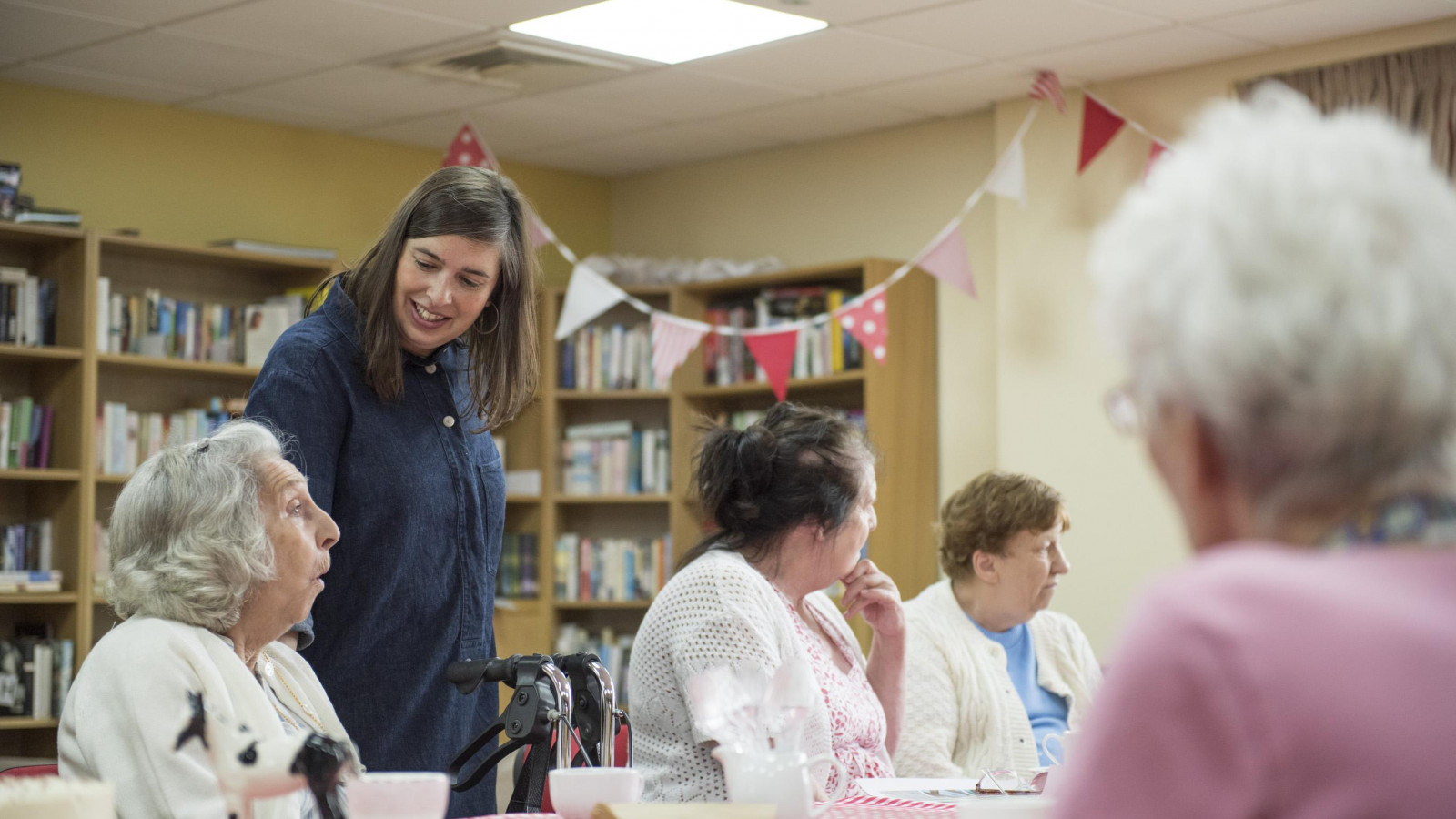 Sheila Ghelani stands at a table decorated with table cloths and teacups. In the background is some red and white bunting. Sheila is smiling and talking to an older woman who is sat at the table. People sit around the table and chat.
