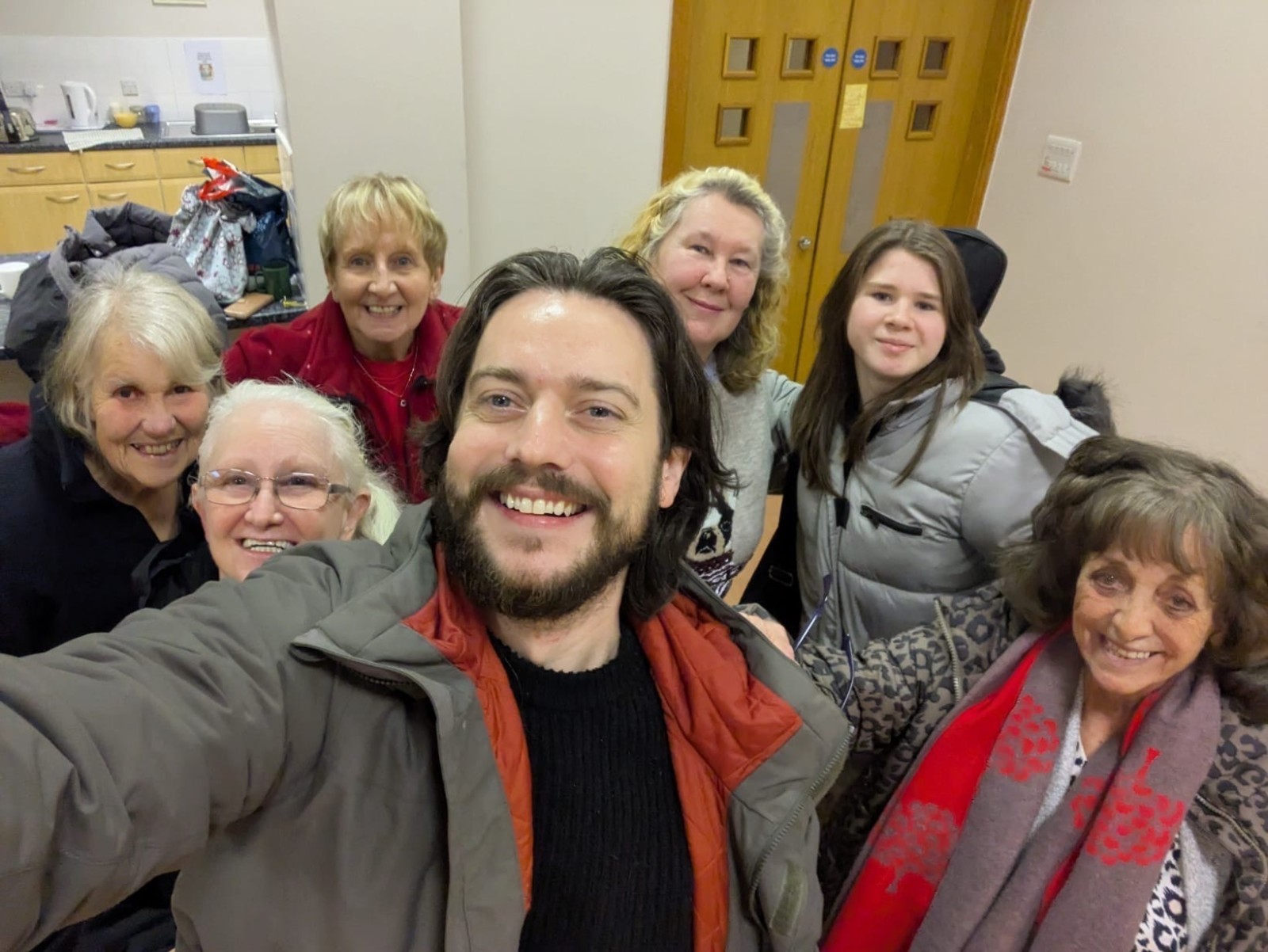 A group of 6 women and 1 man stand facing the camera smiling. The man, who has dark hair and facial has his arm outstretched holding the camera to take a group selfie.