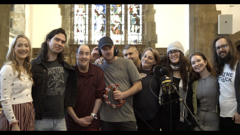 A group of nine people stand in a line in front of a microphone, they are all smiling. One person holds a tambourine.