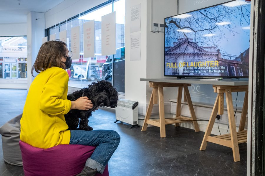 A woman in a yellow jumper holds a small fluffy black dog as she sits and watches a film in the gallery.