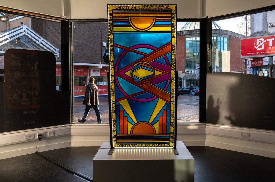 Photo of an art work by Sophie Mahon: A standalone stained glass window is displayed on a plinth. It contains shapes, images and words to do with St Helens.