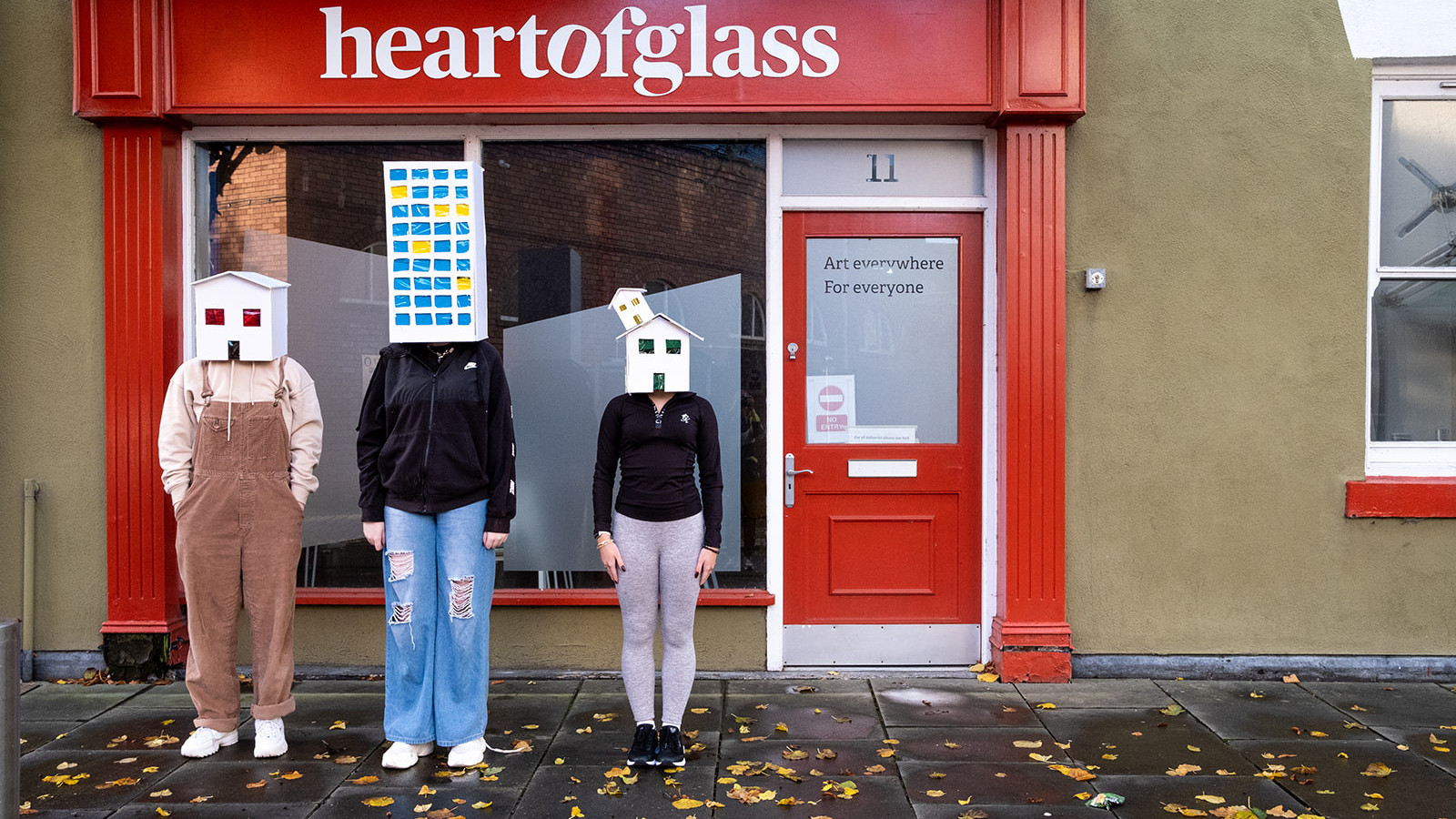 Three people stand side by side. They each wear a paper house over their head. You cannot see their faces.