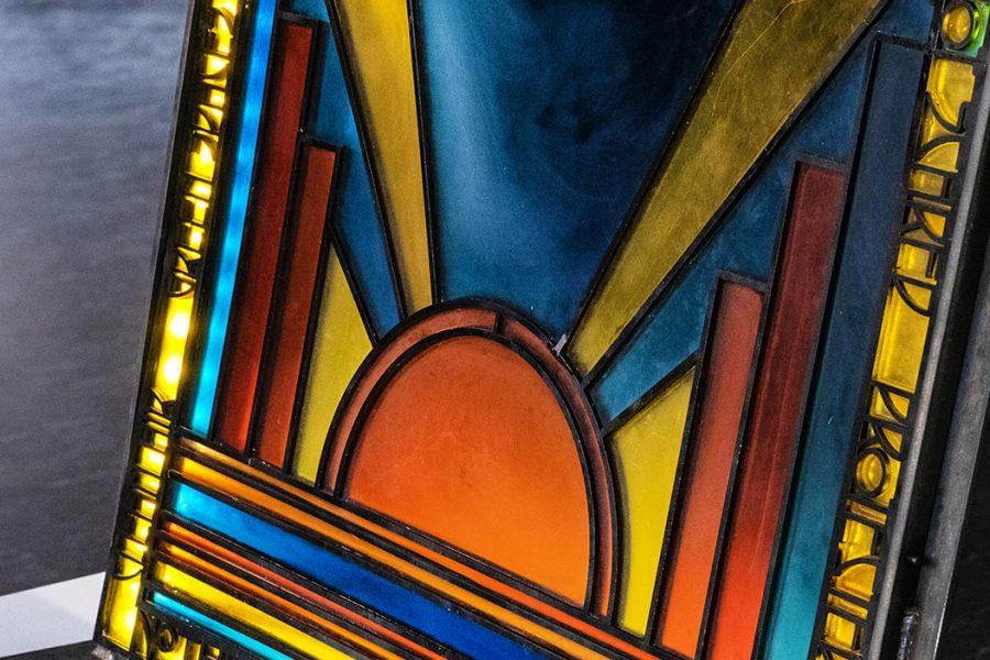 The bottom section of a colourful stained glass panel stands in a shadowy room. Sunlight illuminates sections of the colourful glass.