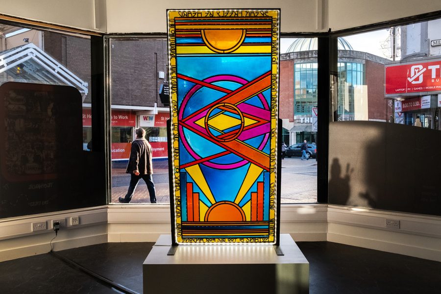A large stained glass panel stands tall on a grey/white plinth. The panel stands in a room surrounded by large windows and the light shines through the colourful panel, illuminating it's pattern.