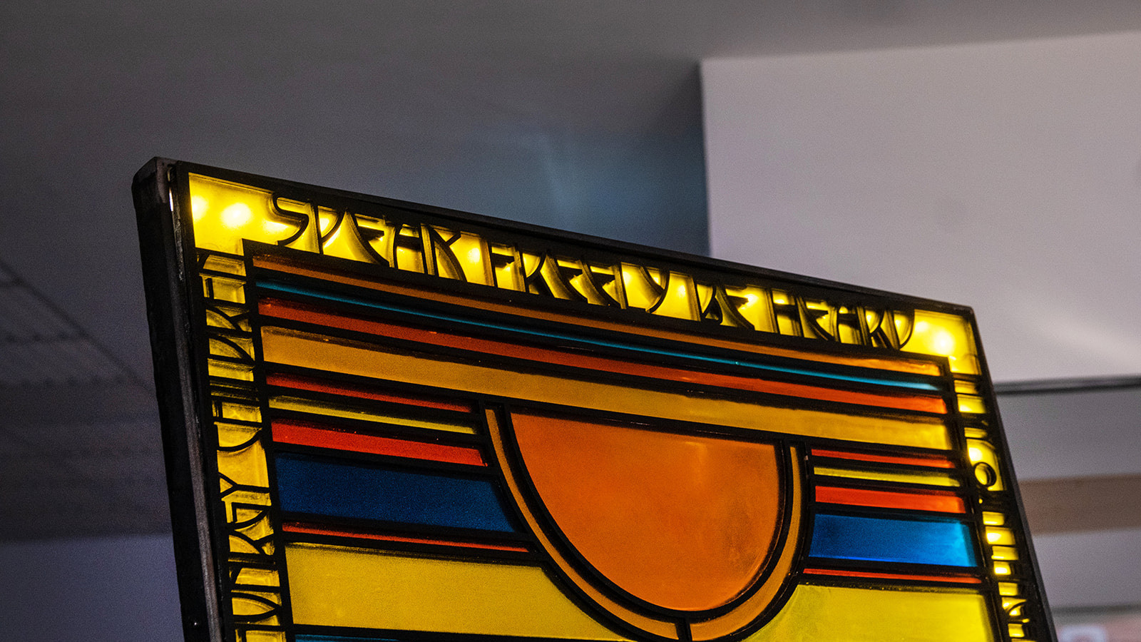 The top section of a colourful stained glass panel stands in a shadowy room. Sunlight illuminates a section of the panel which reads 'Speak Freely Be Heard' in black text on a yellow background.