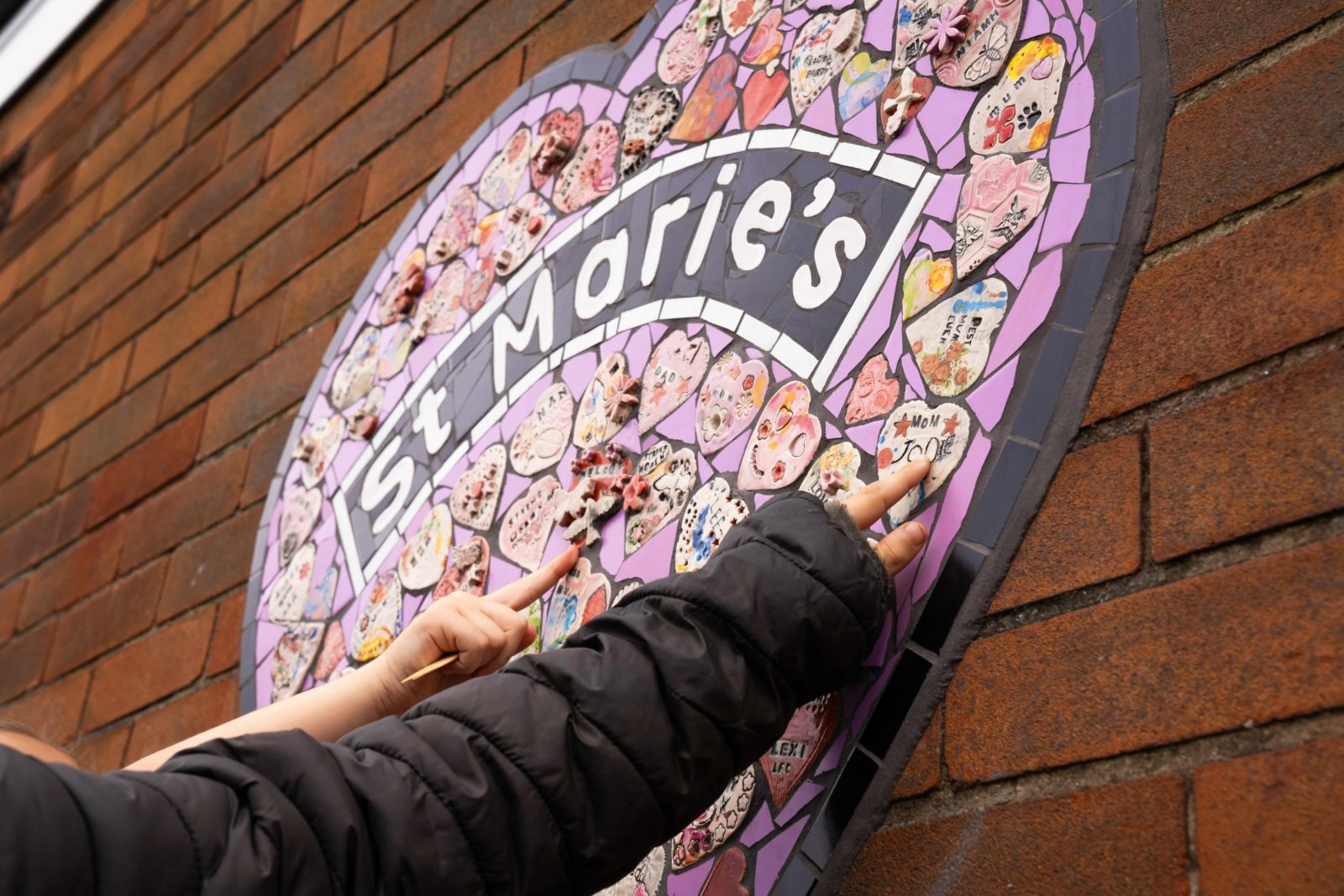 2 children’s hands reach up and point at 2 hearts on the mosaic.
