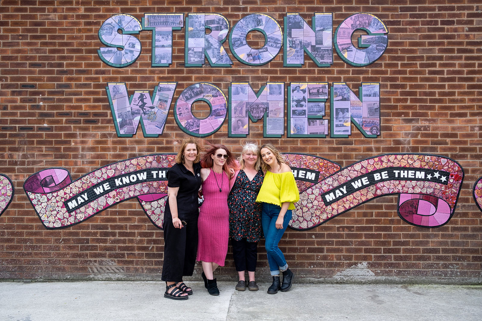 Heart of Glass producers Anna and Rhyannon, Artist Carrie Reichardt and Mentee Claire Eddleston smile for the camera with arms around each other in front of the mural.