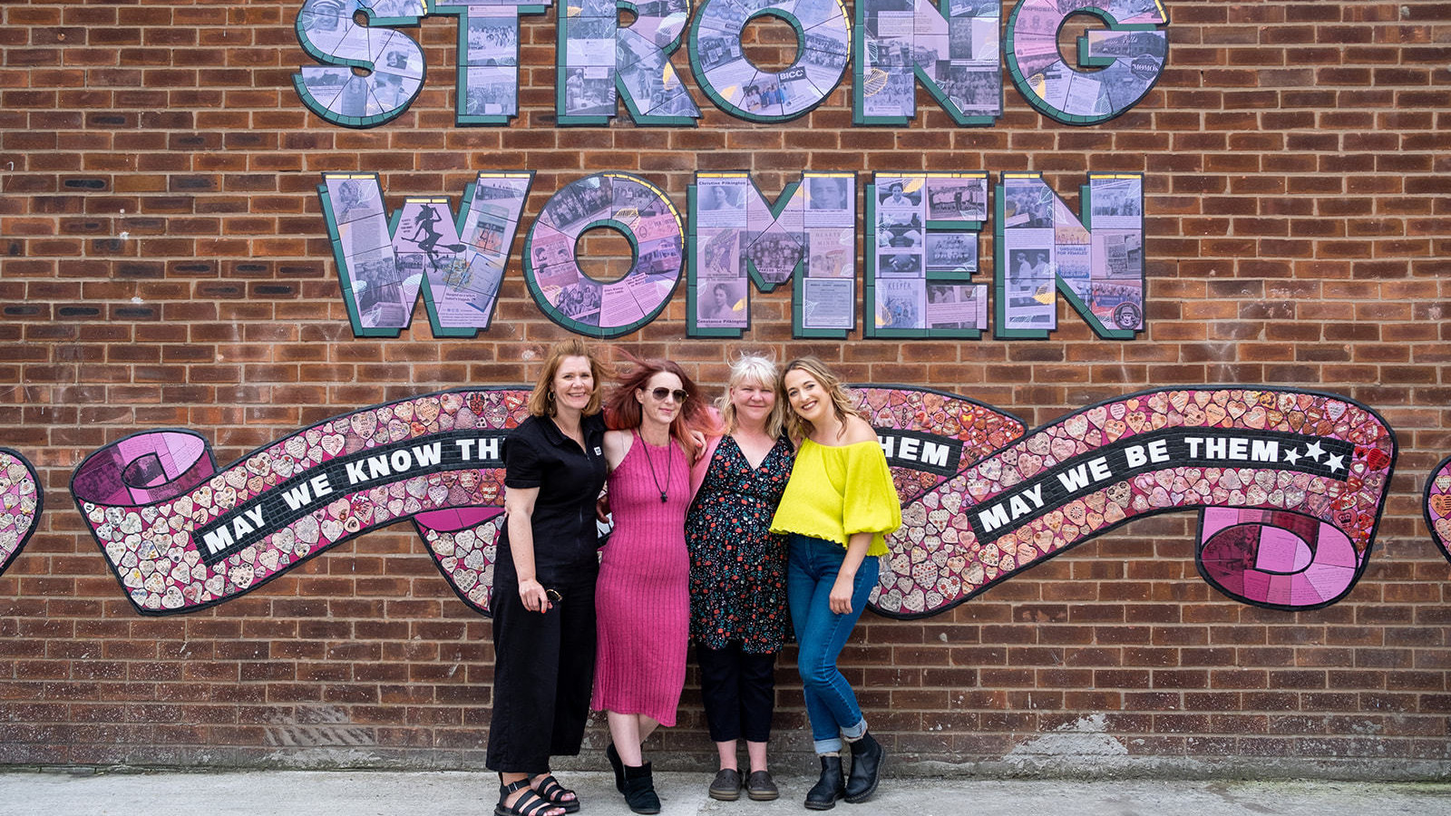 Heart of Glass producers Anna and Rhyannon, Artist Carrie Reichardt and Mentee Claire Eddleston smile for the camera with arms around each other in front of the mural.