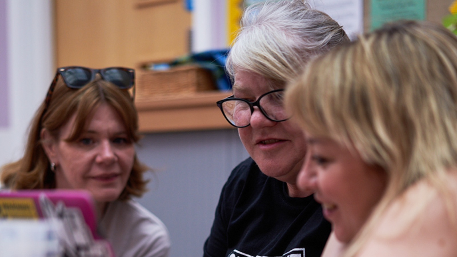 Three women sit around a laptop, looking at the screen and smiling