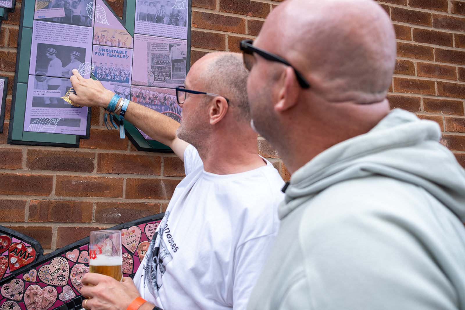 Two men point at the tiles on the Strong Women of St Helens mural, they are printed with historic photos and text.