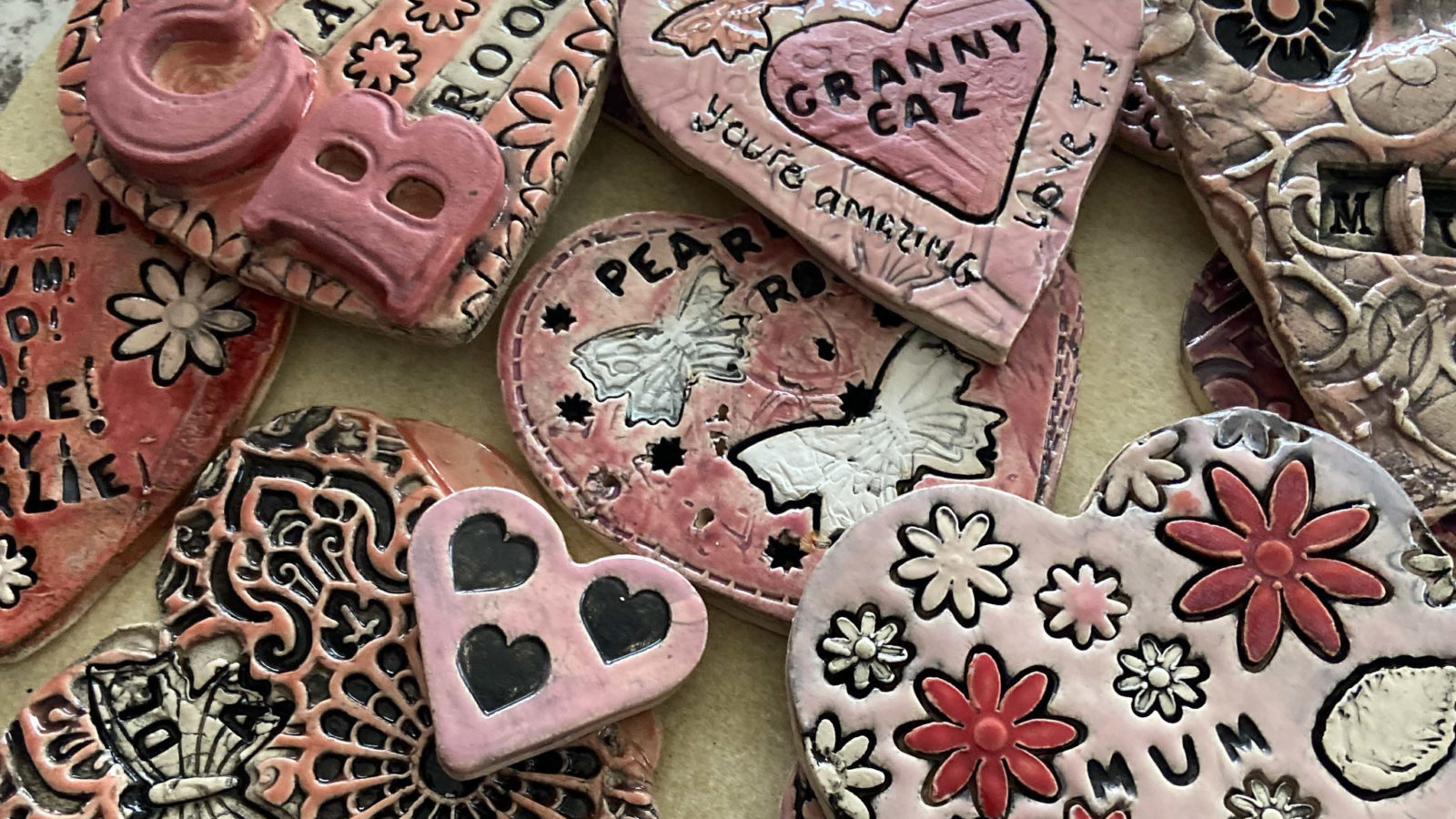 A selection of glazed clay hearts in varieties of pinks and reds, with engravings and writing on them, are laid out on a surface.