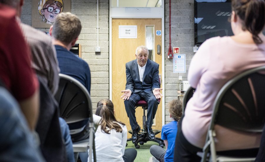 A man in a suit, with light, short hair sits on a stool in front of an audience. He gestures with his hands and is talking to children sat on the floor.