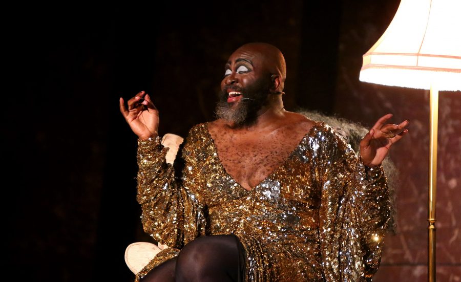 A person with a dark beard wears a gold, glittering gown and wits in a darkened room next to a lamp. They are singing.