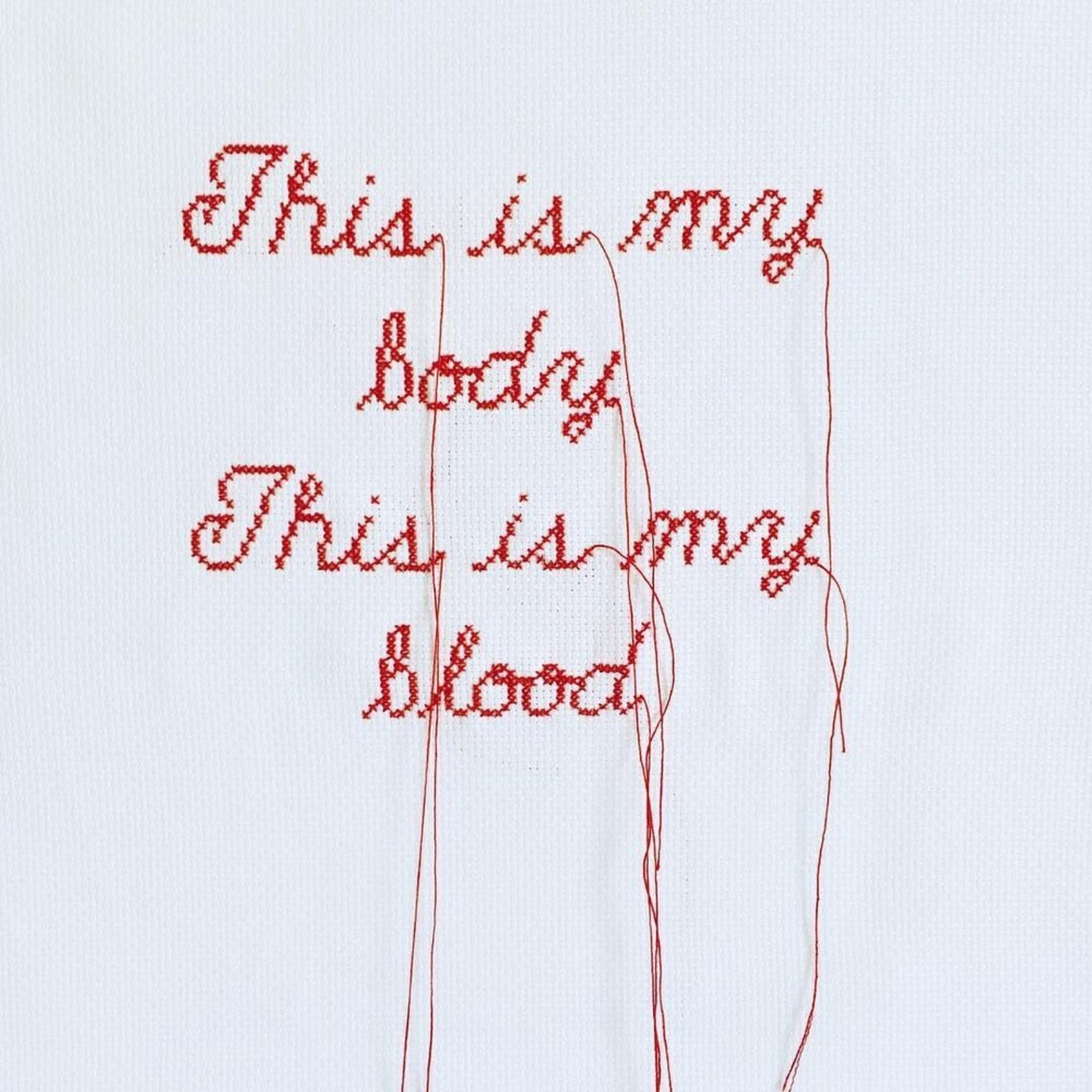 Red letters cross stitched on to white fabric. The letters read 'This is my body. This is my blood'. Red cotton hangs from the lettering.