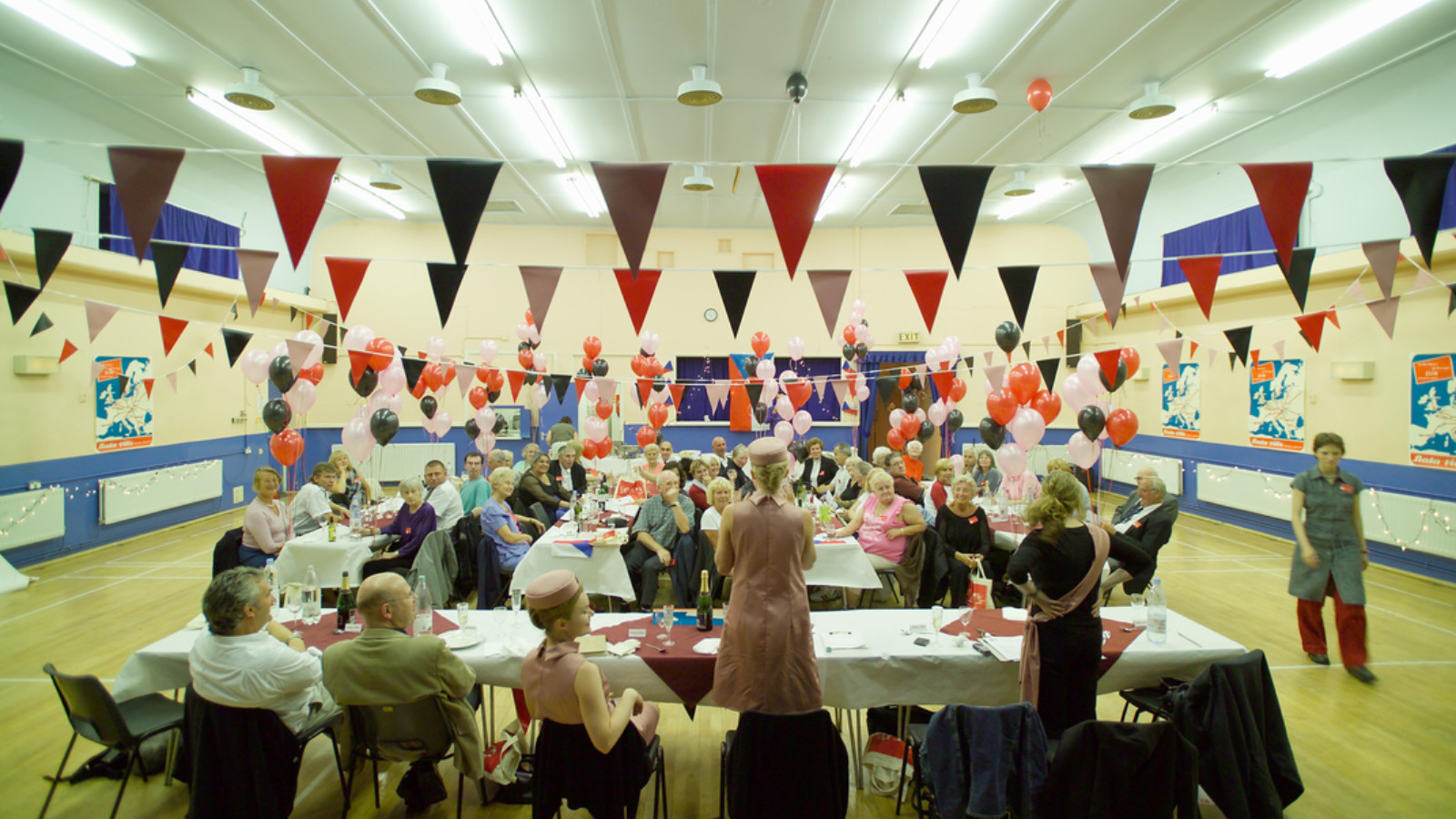 A community hall has tables and chairs set up with colourful bunting and balloons to decorate. People are sat at the tables watching a woman in a pink hat and dress speak.